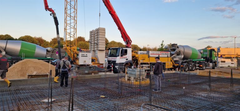 Betonpumpe und zwei Fahrmischer an einer bewehrten Bodenplatte, Arbeiter auf der Bewehrung