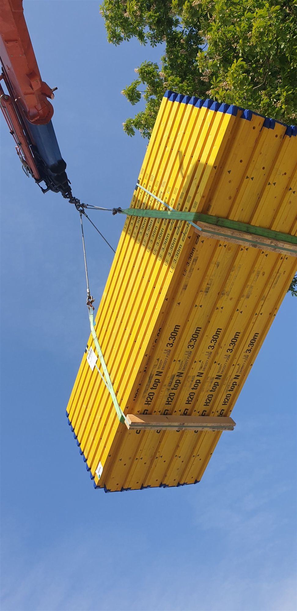 Ein gebündeltes Paket gelber Schalungsträger hängt am Kranhaken vor blauem Himmel